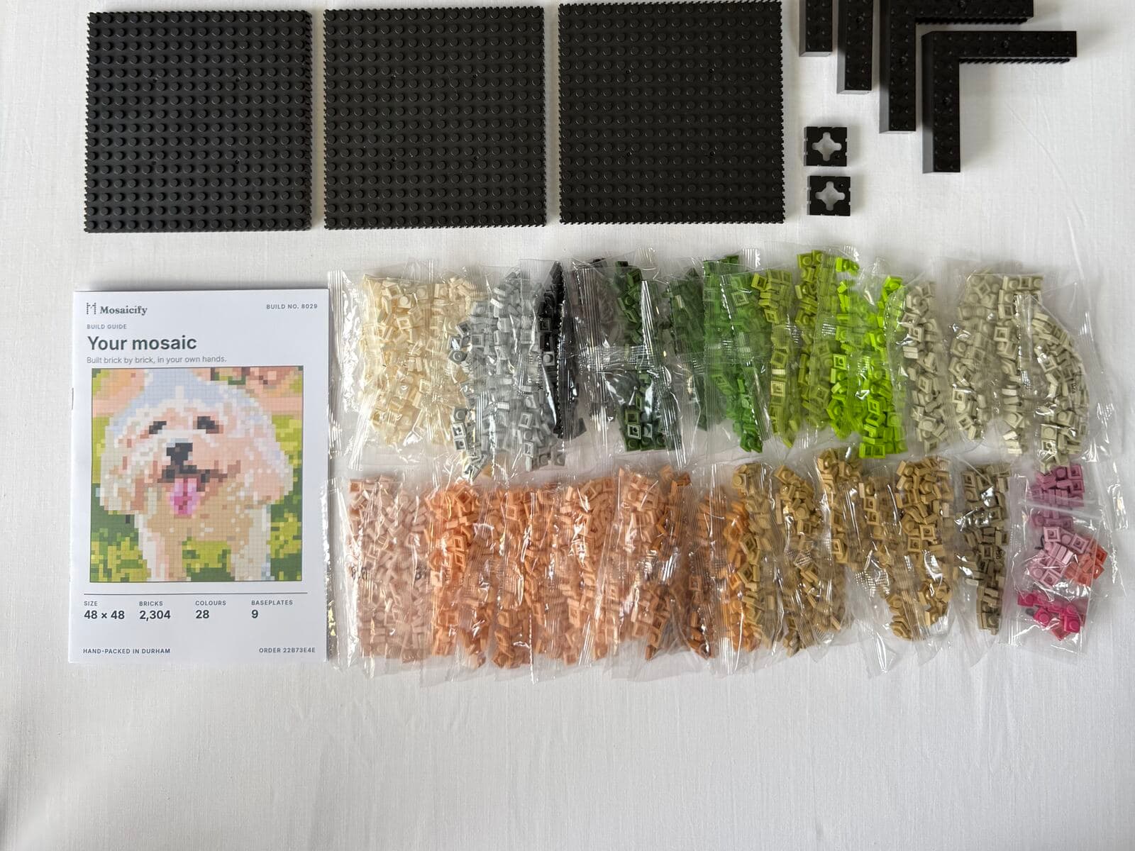 A Mosaicify kit laid out as it's hand-packed in the Durham workshop: colour-sorted brick bags, black baseplates, frame parts and the printed build booklet